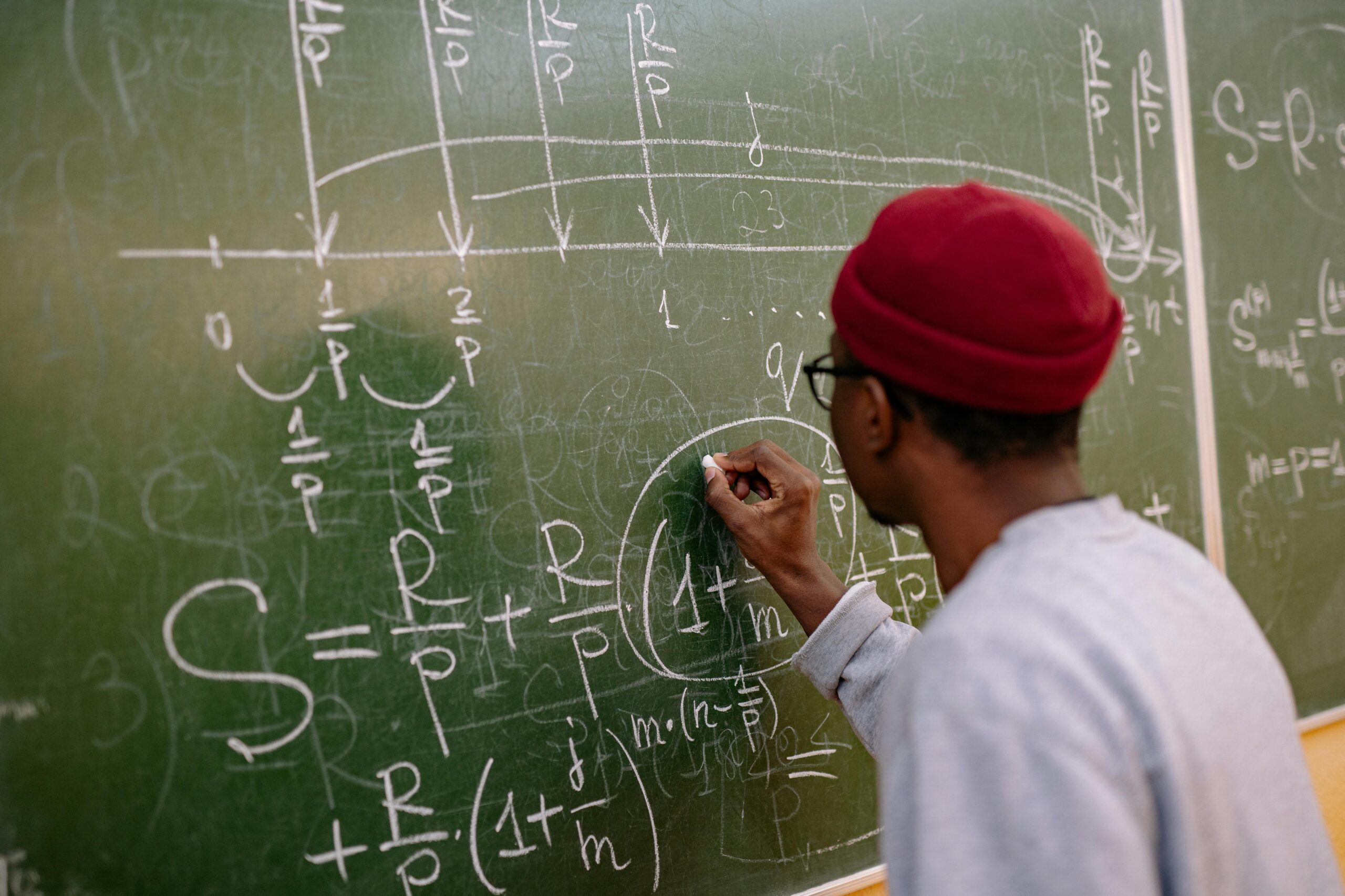 Ana Sayfa Student writing complex mathematical equations on a chalkboard in a classroom setting.